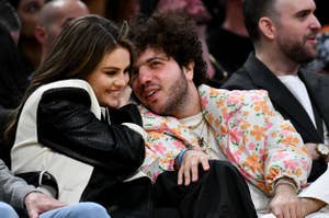 Selena Gomez and The Weeknd sitting close, Selena leans on him, both smiling, at a public event. He wears a floral jacket