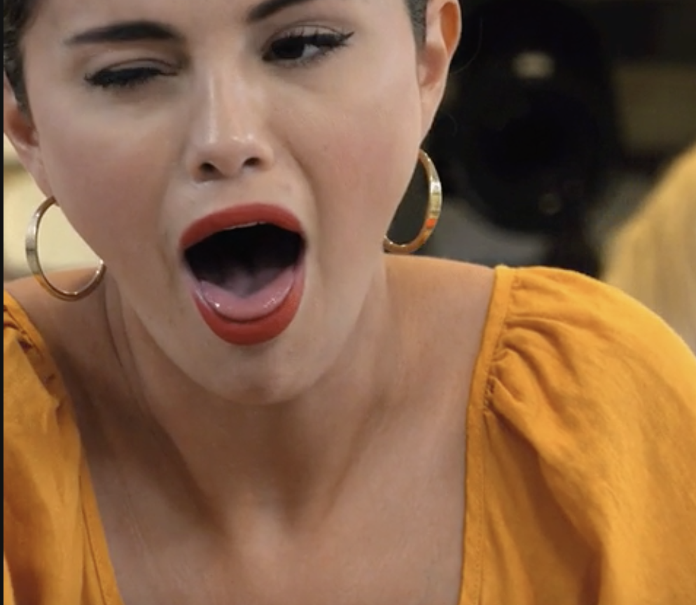 A person with hoop earrings and a yellow top appears surprised with mouth open wide
