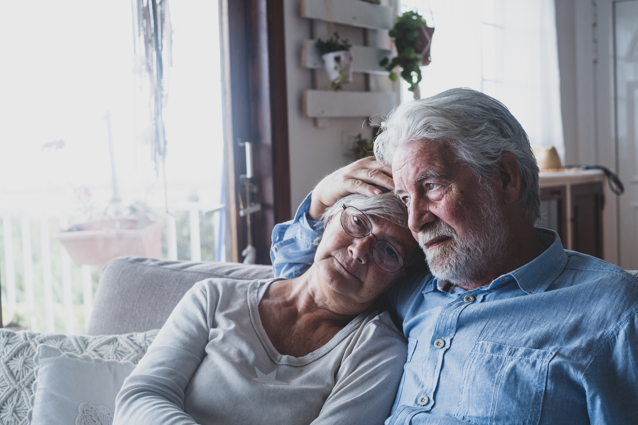 Elderly couple embracing and looking pensively in the distance, seated inside a home