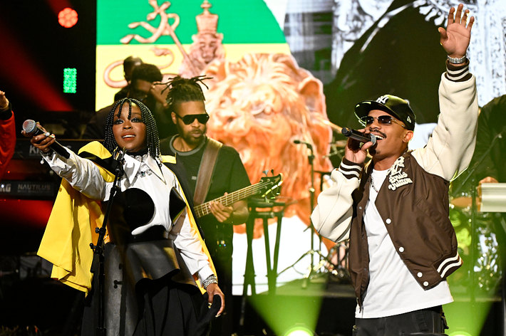 Two musicians perform on stage with a band, one in a black cap and jacket, the other with braids and a yellow top