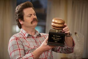 Ron Swanson is shown holding a trophy for a burger cook-off in a plaid shirt