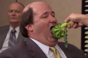 Man in a suit eating broccoli held by another person, with a man in the background looking on