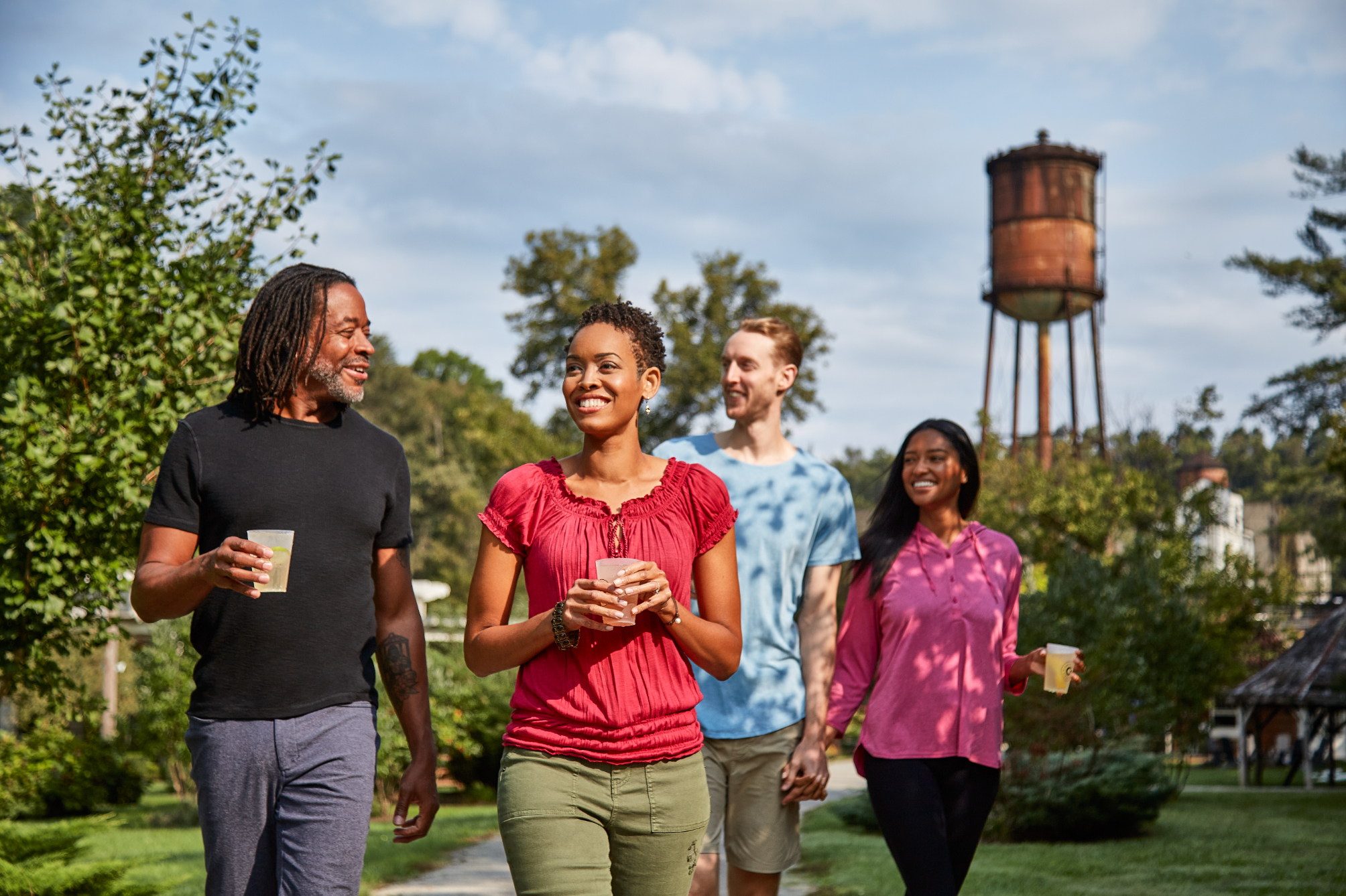 Four friends walking outdoors holding drinks, smiling, with a water tower in the background