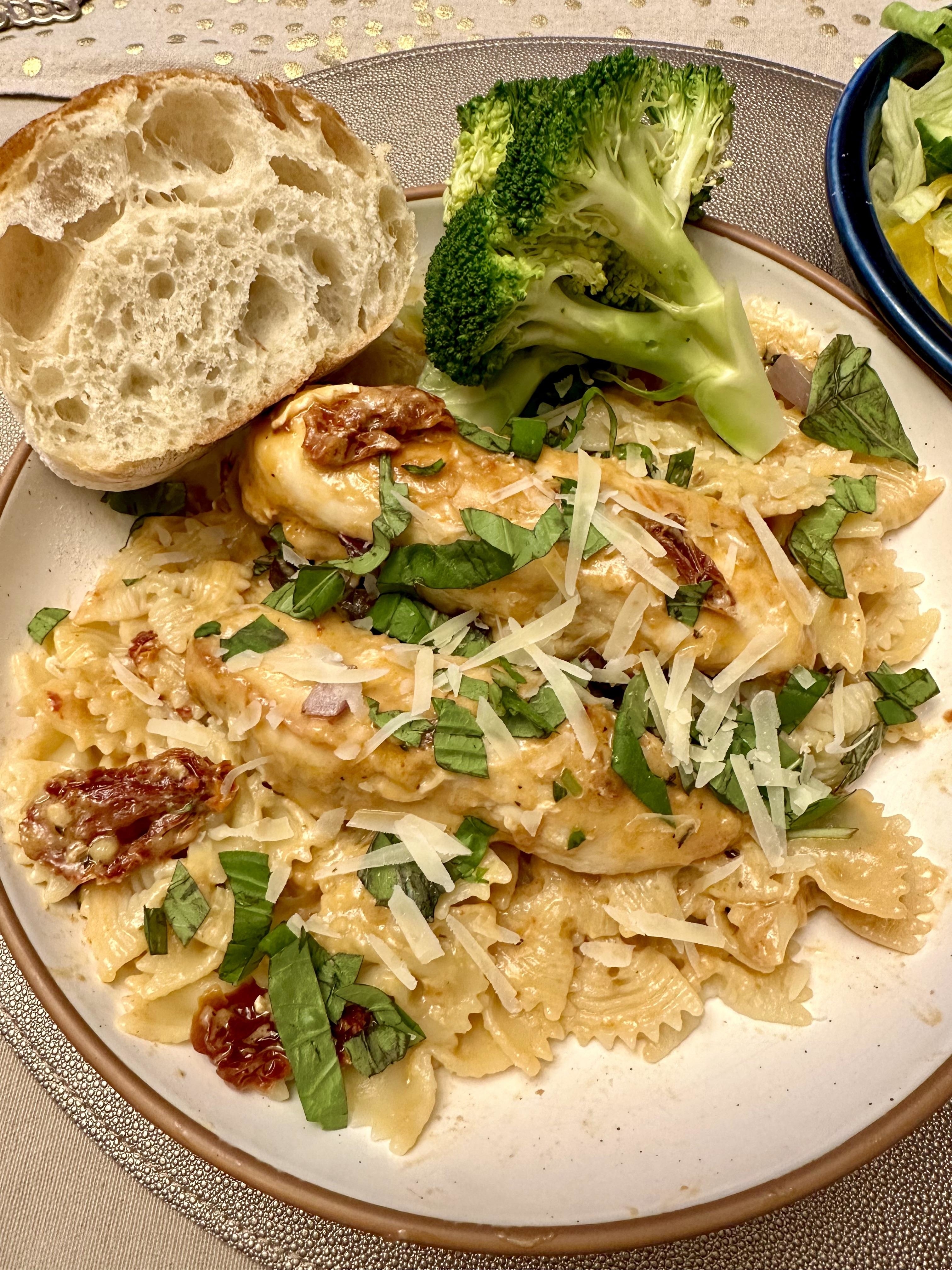 Plate with pasta, chicken, sun-dried tomatoes, broccoli, and topped with shredded cheese and basil