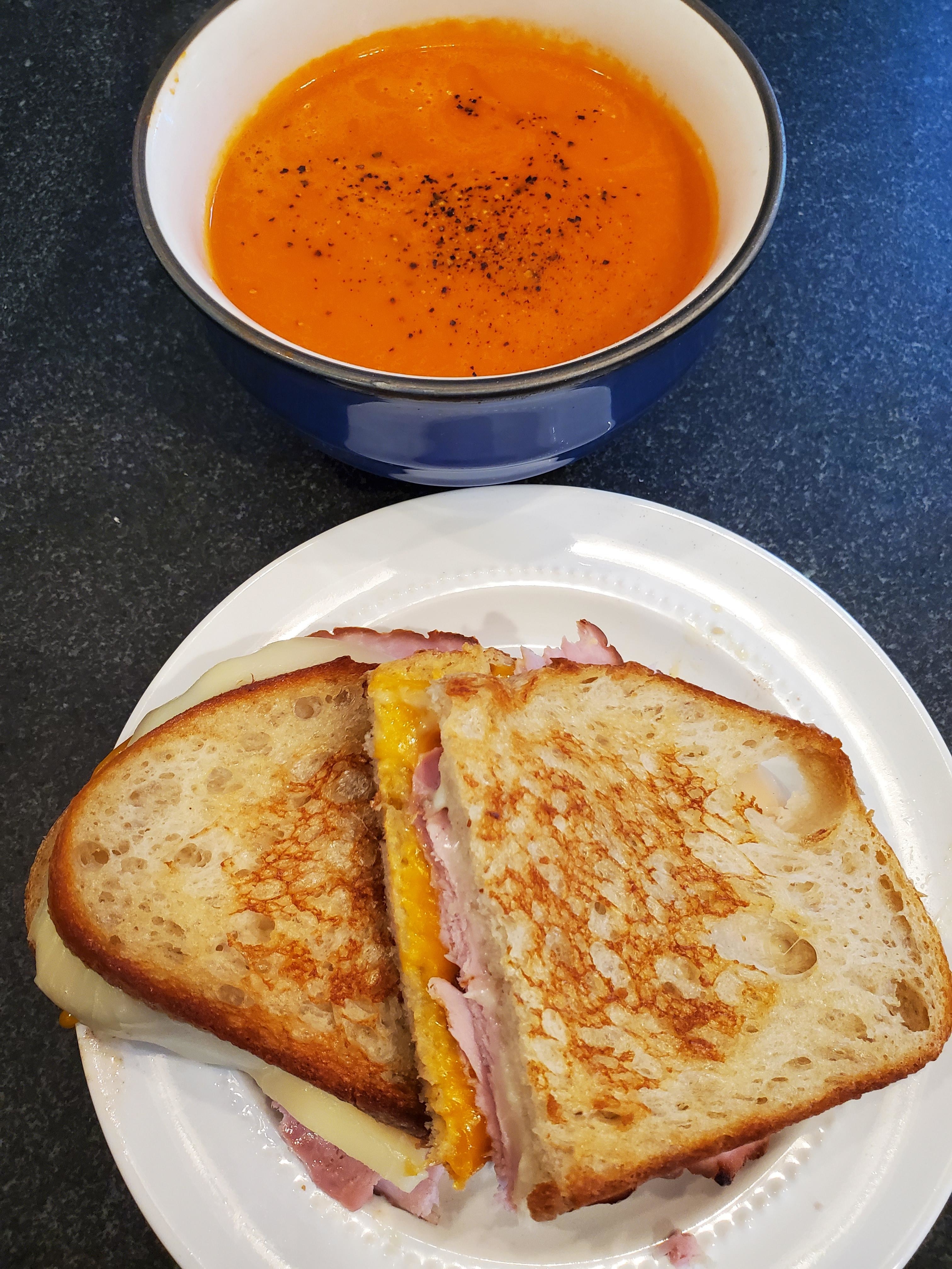 A bowl of tomato soup next to a grilled cheese sandwich with ham on a plate