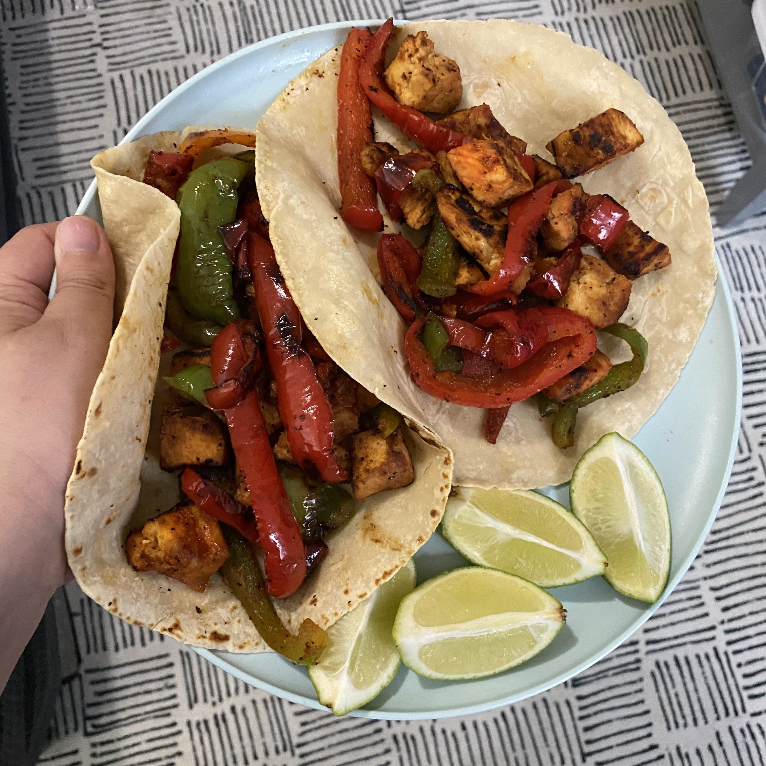 Hand holding two tacos filled with grilled chicken and bell peppers, with lime wedges on the side