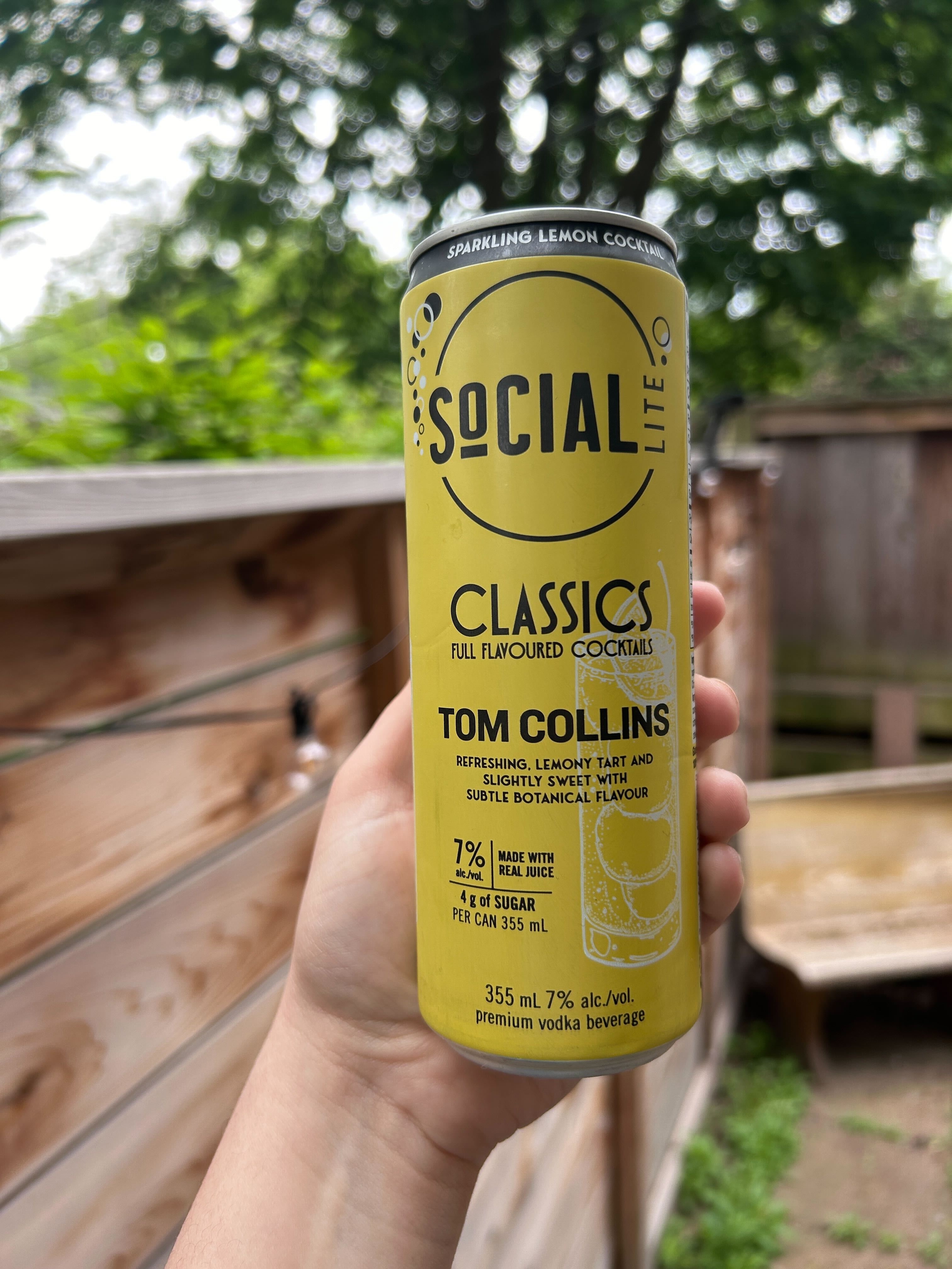 Hand holding a can of Social Lite Sparkling Lemon Cucumber Tom Collins cocktail outdoors
