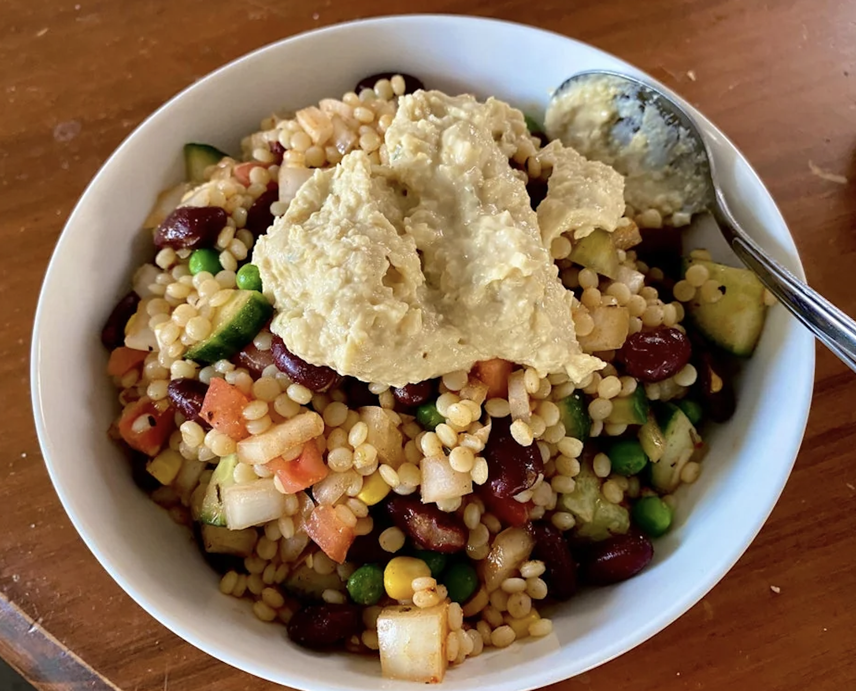 A bowl of mixed grain salad with a dollop of hummus on top, next to a spoon