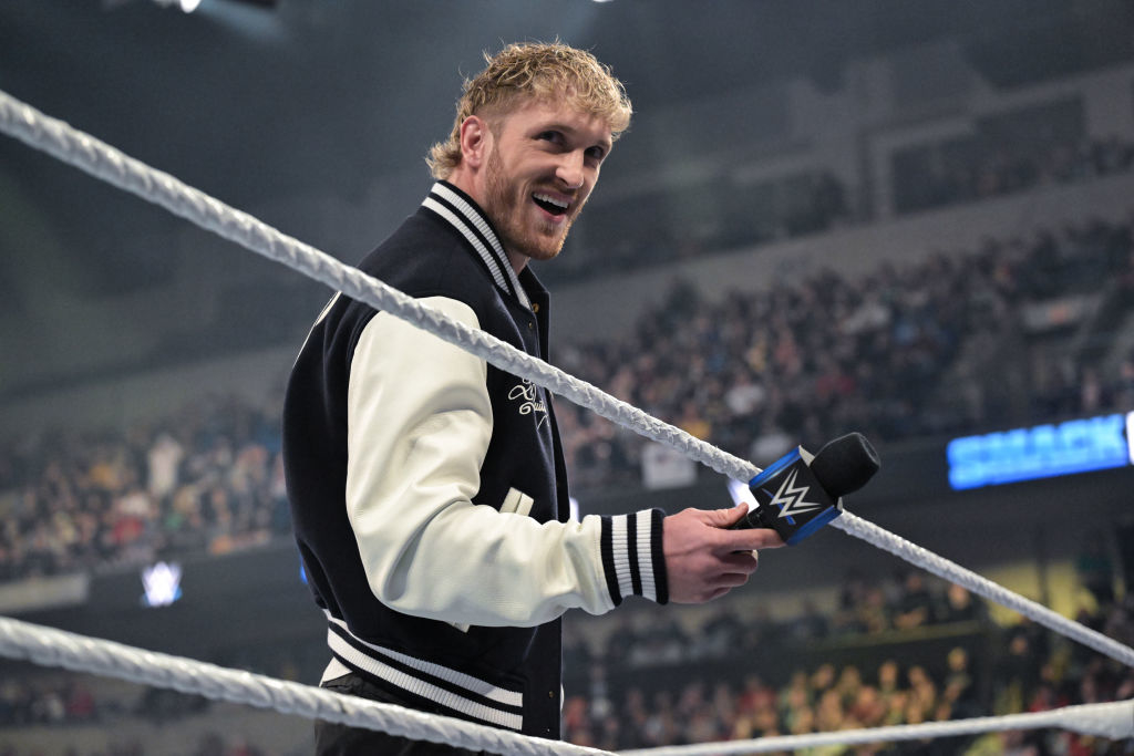 ogan in a ring holding a microphone, wearing a varsity jacket, with an audience in the background
