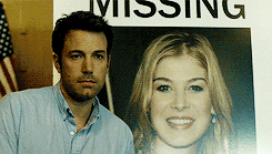 Ben Affleck smiles next to a "MISSING" poster of Rosamund Pike from the movie "Gone Girl."