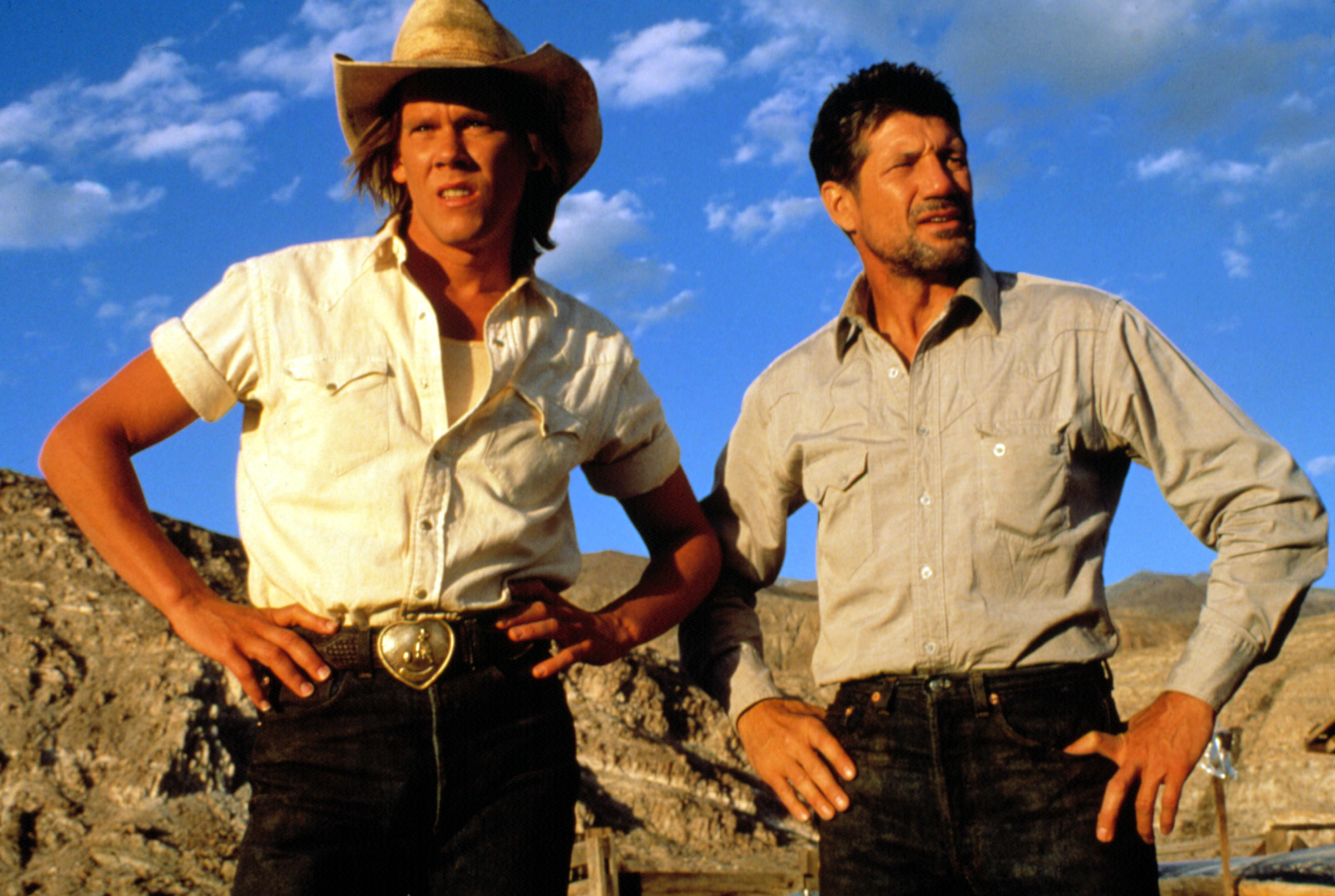 Two actors from a movie pose, one in a cowboy hat and belt buckle, the other in a casual shirt, both looking afar