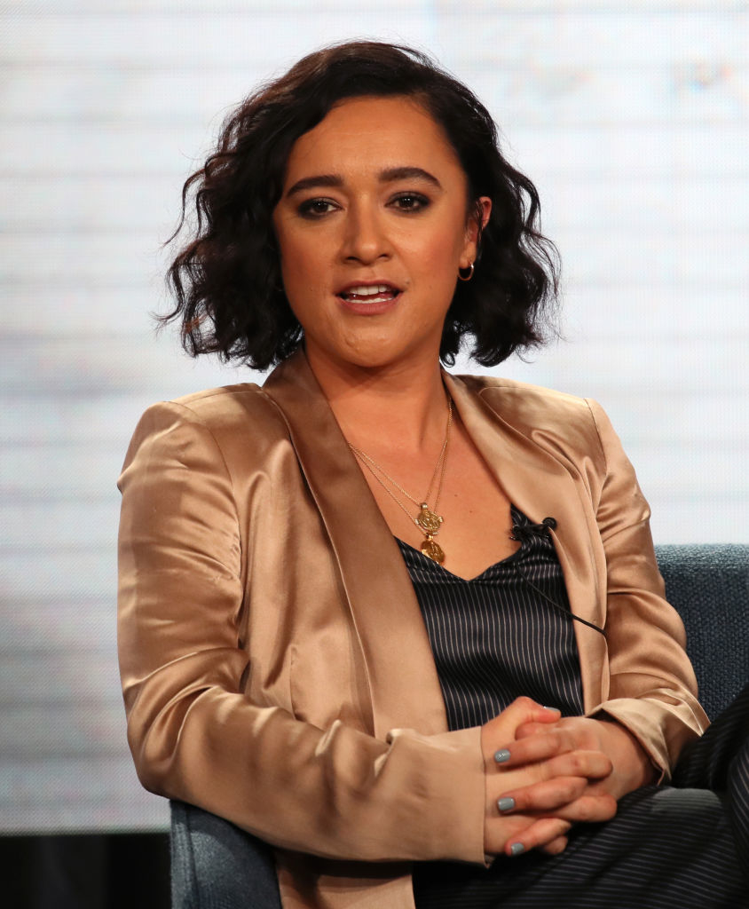 Woman in a satin blazer over a top, sitting with crossed hands, looking to the side, expression attentive