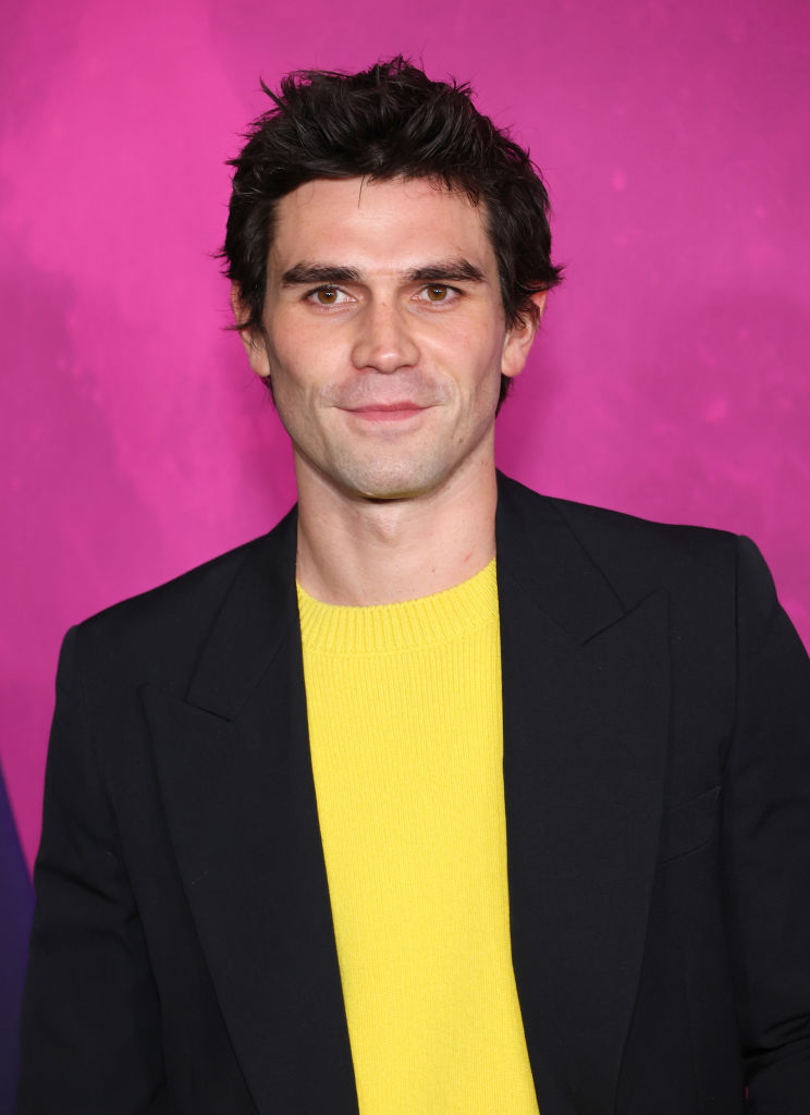Man in a black blazer over a yellow sweater, smiling against a pink background