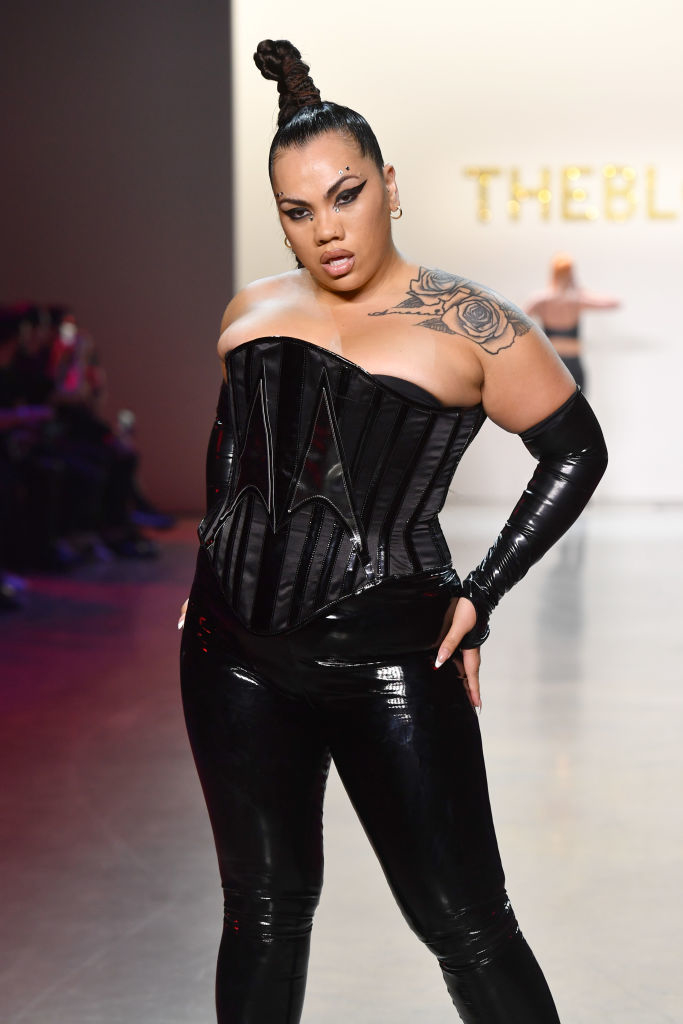 Person stands confidently in a black corset top and shiny pants with a high ponytail and tattoo on arm