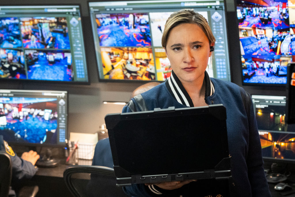Character in a control room looking intently at the camera, surrounded by screens showing surveillance footage
