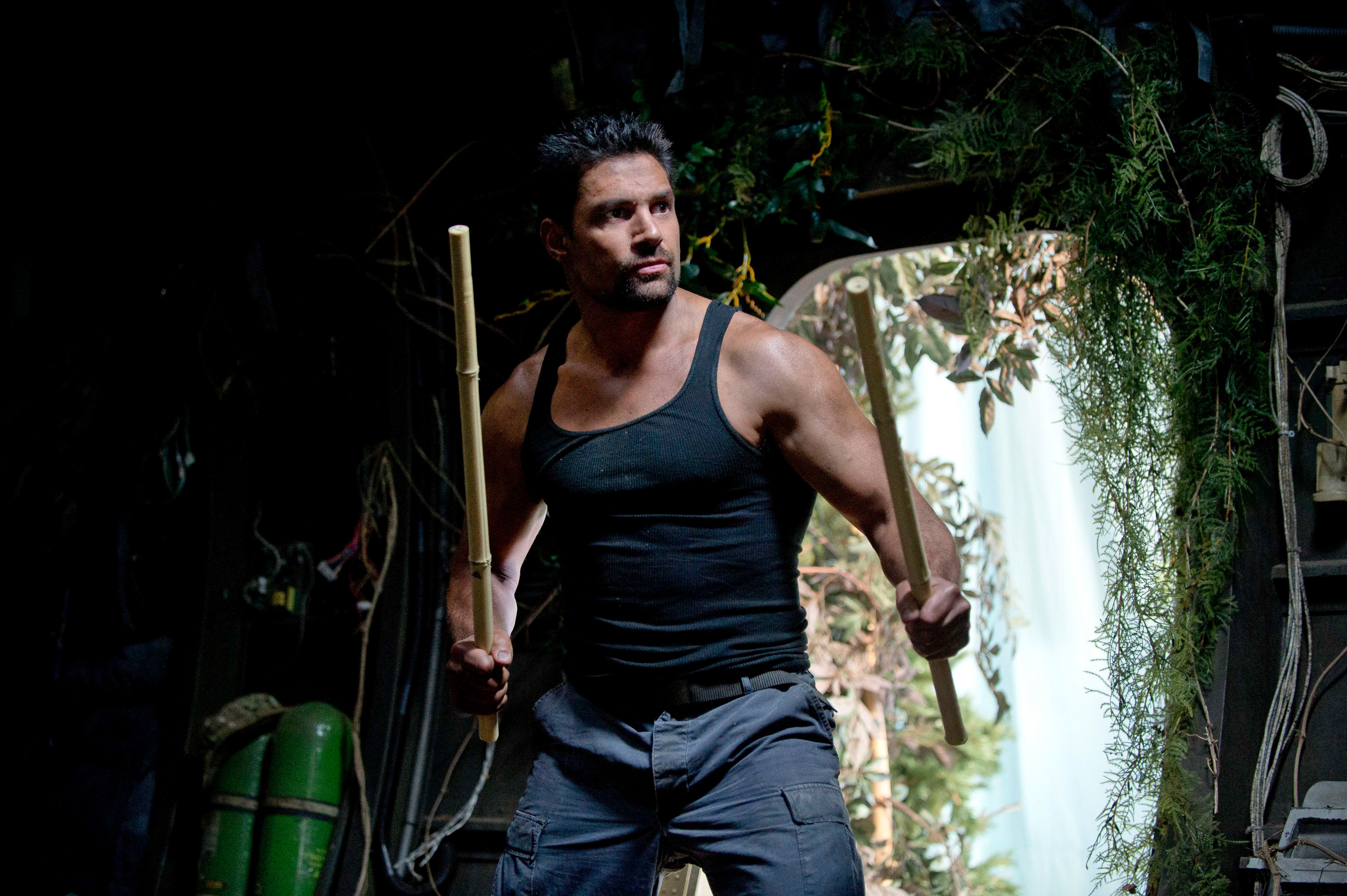 Man carrying wooden staff, wearing tank top and cargo pants, intense expression