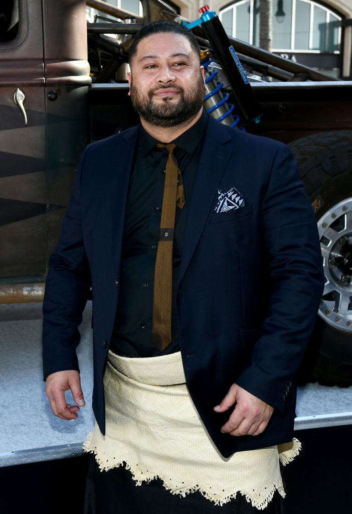 Man in a suit jacket and traditional Polynesian skirt poses confidently
