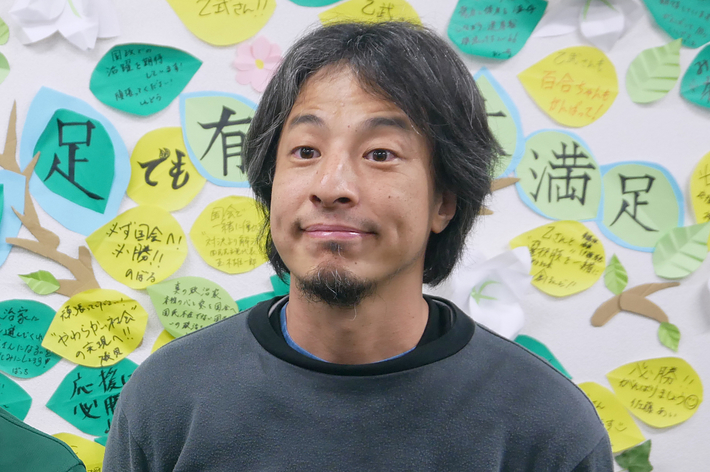 二人の男性がカメラに向かって笑顔で立っており、背景には沢山のメッセージが書かれた紙が壁に貼られている。