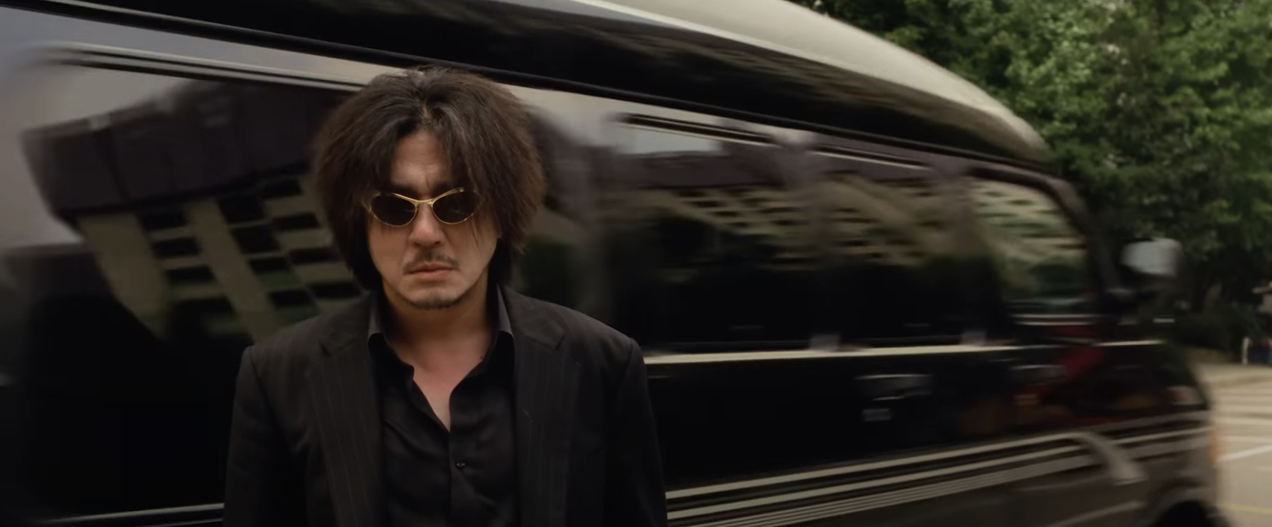 Man in black suit and sunglasses walks in front of a vehicle, reminiscent of an intense movie scene