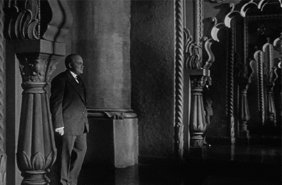 Actor in a classic film standing solemnly by a column with ornate details, in a dark, moody scene