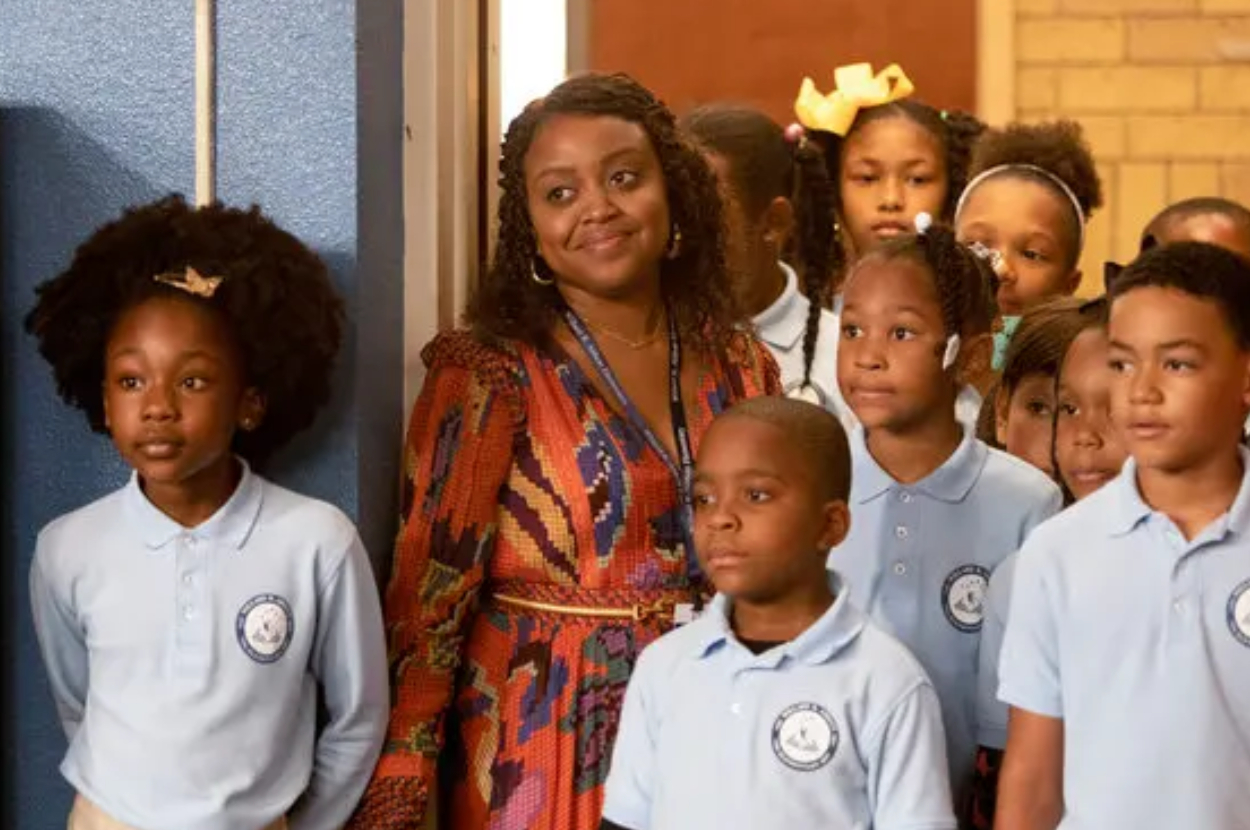 A group of young students in uniform with a smiling woman wearing a patterned dress standing in a school hallway