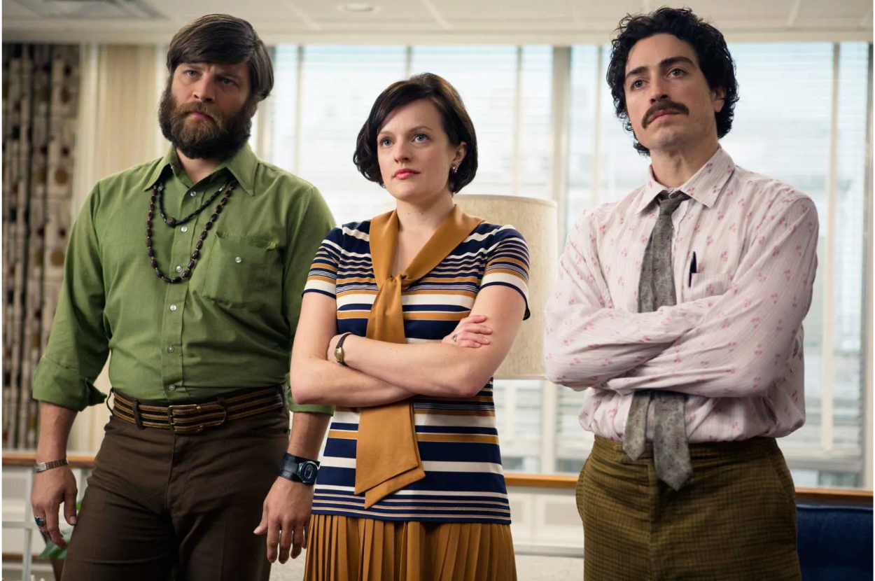 Three actors in 70s attire standing with serious expressions in a room