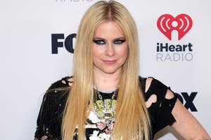 Avril Lavigne posing at an event, wearing a graphic tee, layered necklaces, and a black shredded top