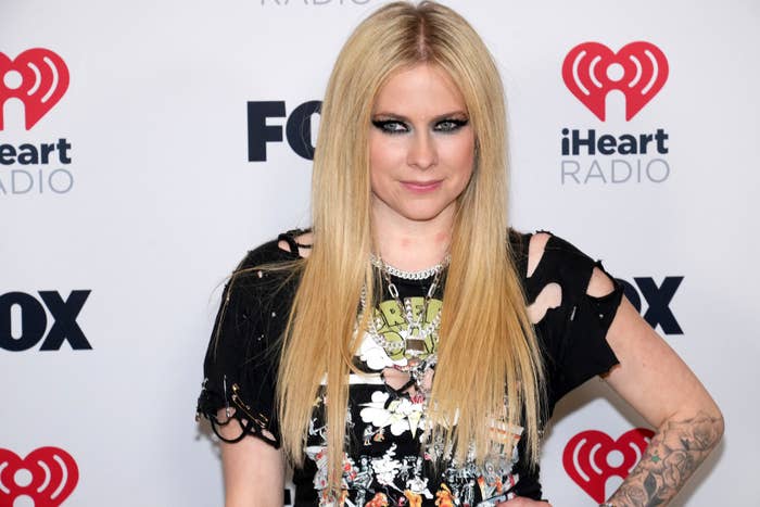 Avril Lavigne posing at an event, wearing a graphic tee, layered necklaces, and a dark shredded top