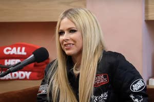 Avril Lavigne in a black jacket with patches, seated, speaking into a microphone