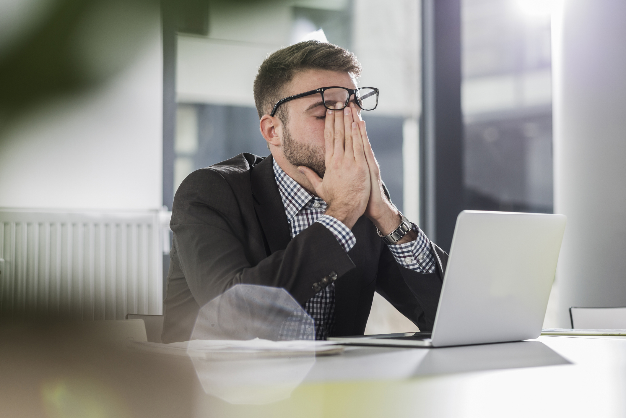 Man in a suit sitting at a laptop appears stressed or tired, covering his face with hands in an office setting