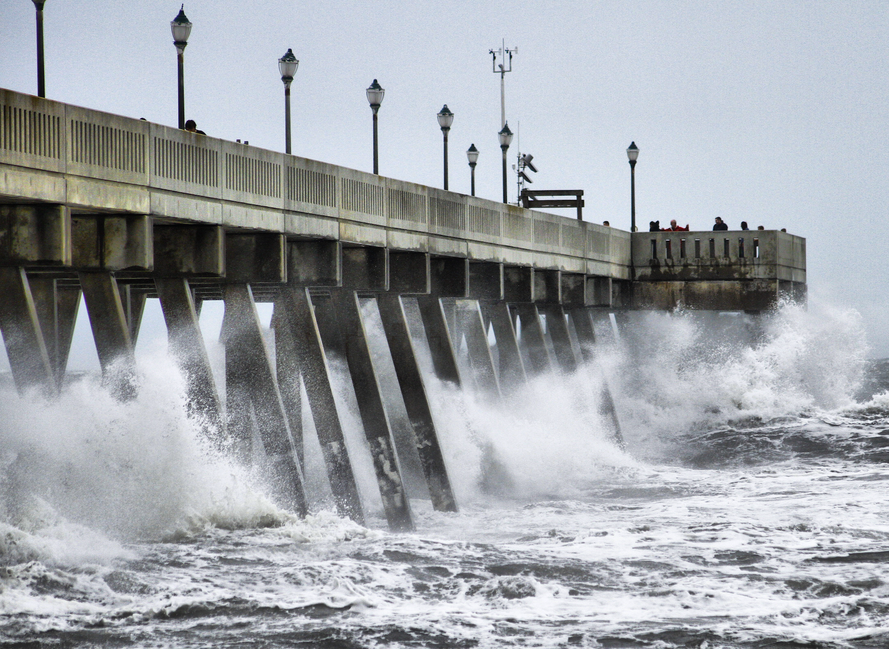 Pier with waves crashing against its pillars, under an overcast sky. People are visible at the far end of the pier