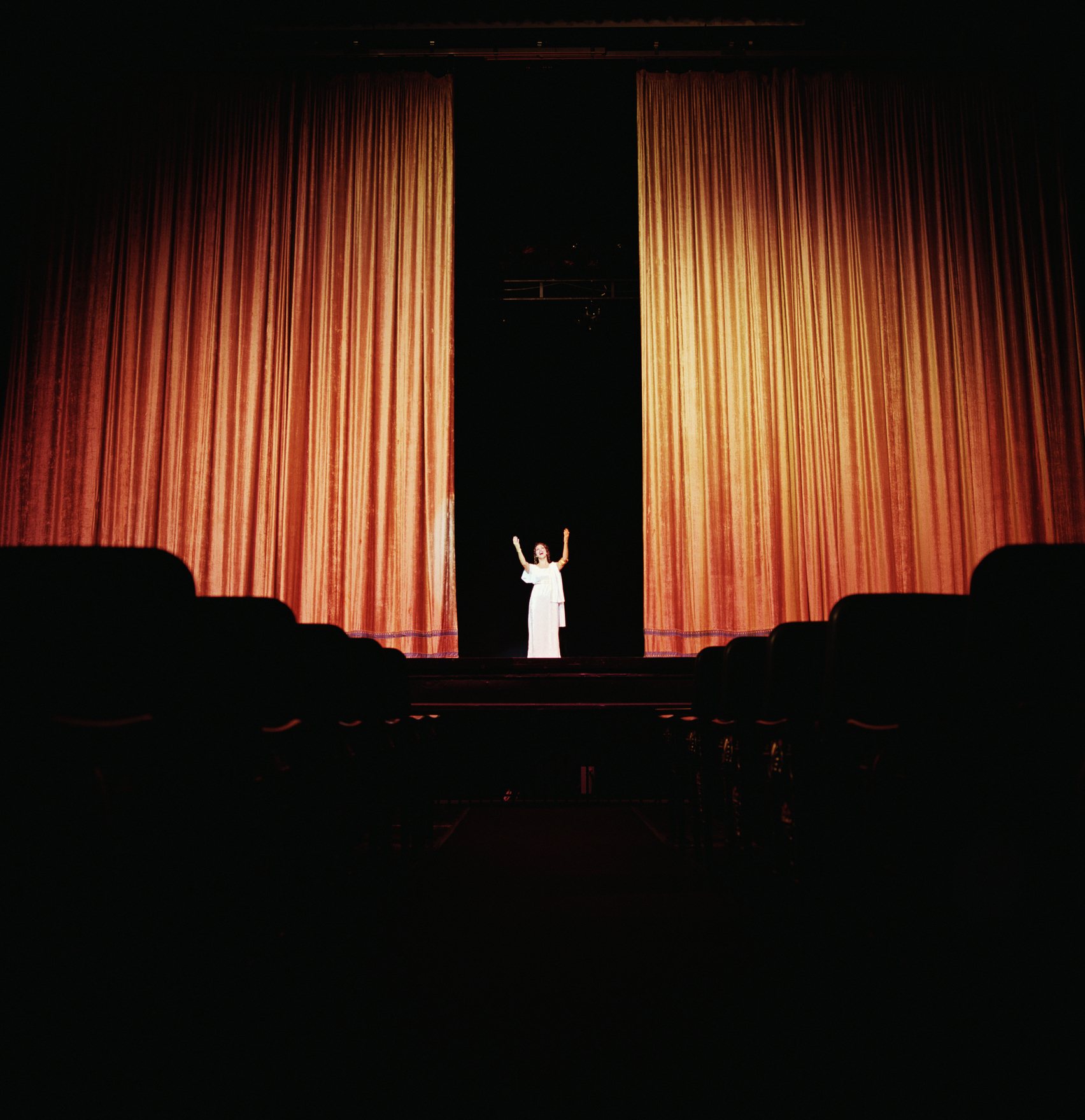 Person on stage with arms raised, audience seats in foreground, theater setting