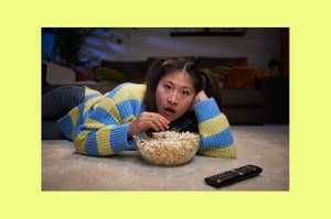 Person lying on stomach watching TV, looking surprised, with a bowl of popcorn and remote nearby