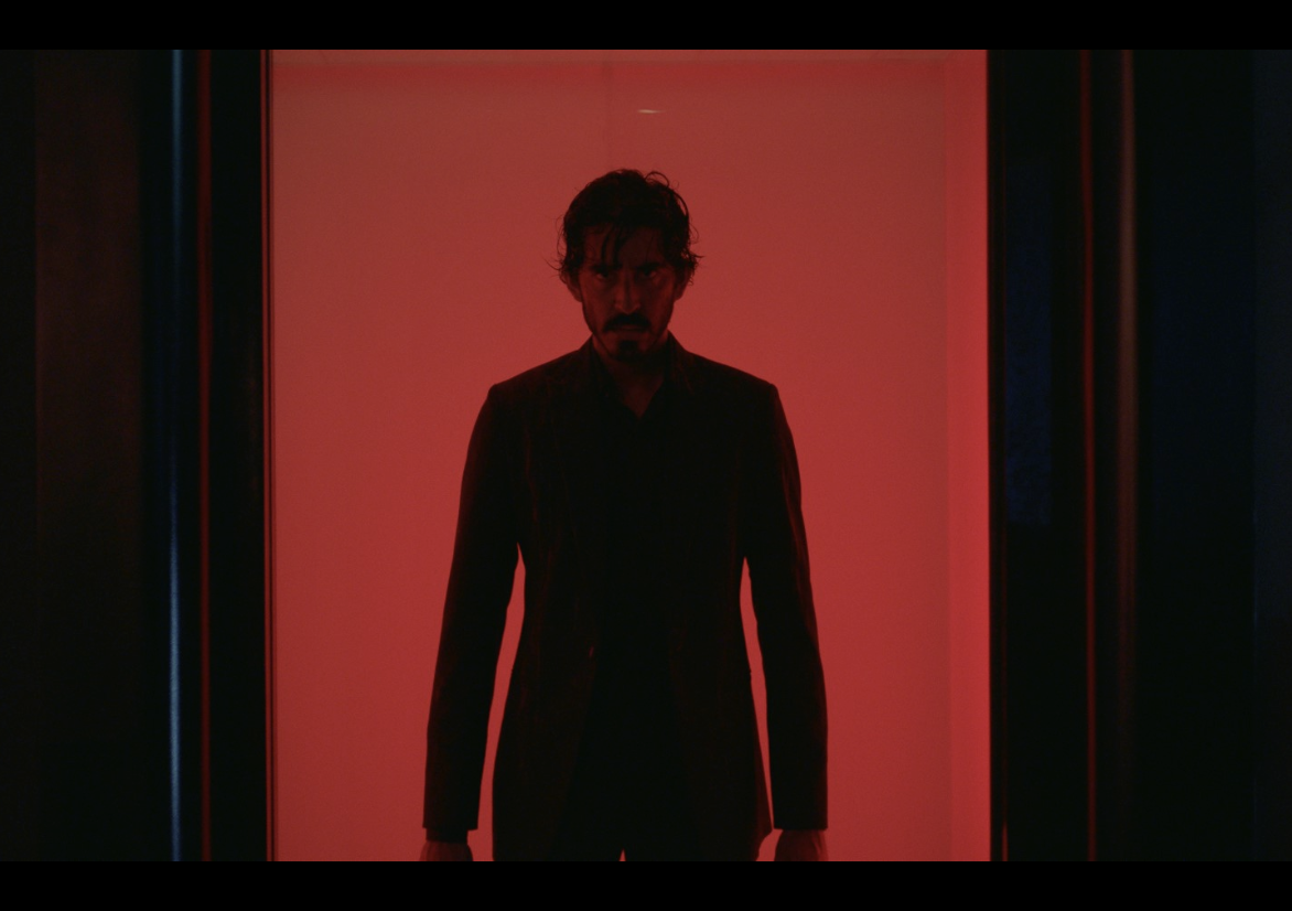 Hombre con traje oscuro, de pie frente a una luz roja de fondo. Escena dramática de serie o película