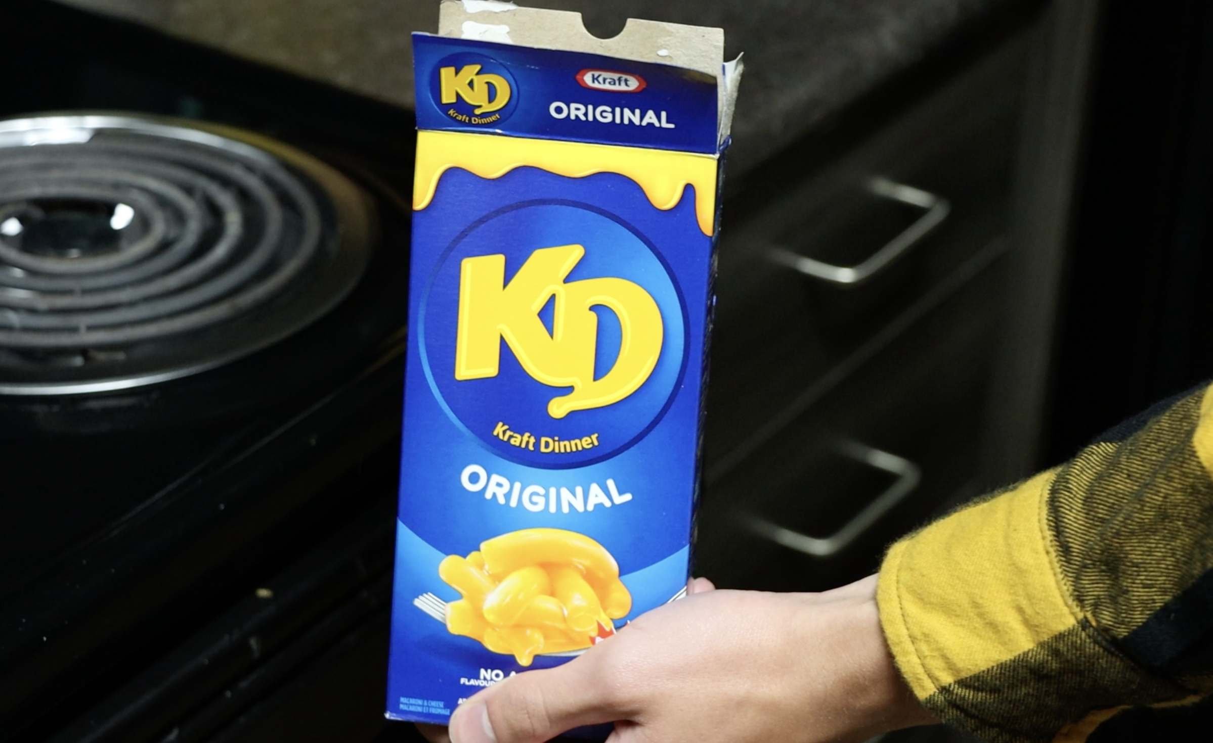 Person holding a box of Kraft Dinner macaroni and cheese in a kitchen setting