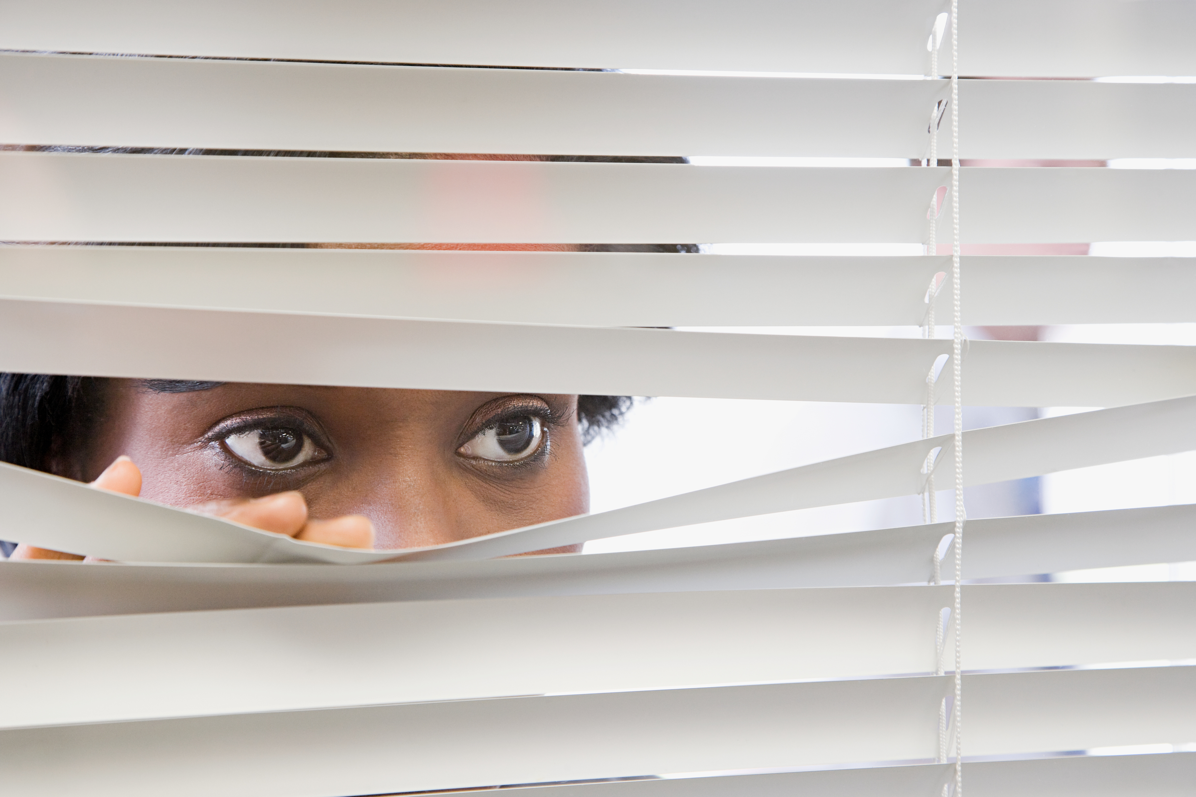 Person peeking through slightly opened blinds, with focus on their eyes, suggesting vigilance or curiosity at work