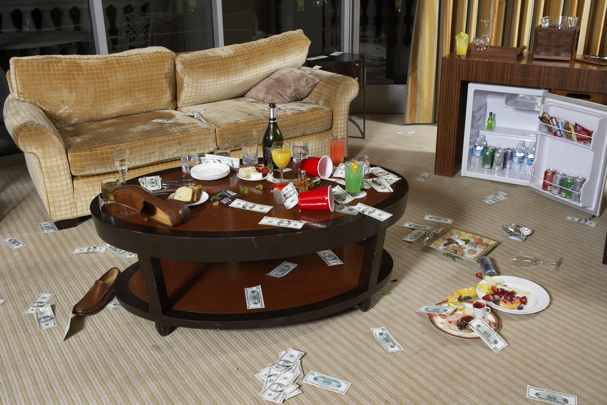 A messy room with scattered money, an overturned chair, beverages and food on the table, and an open mini-fridge