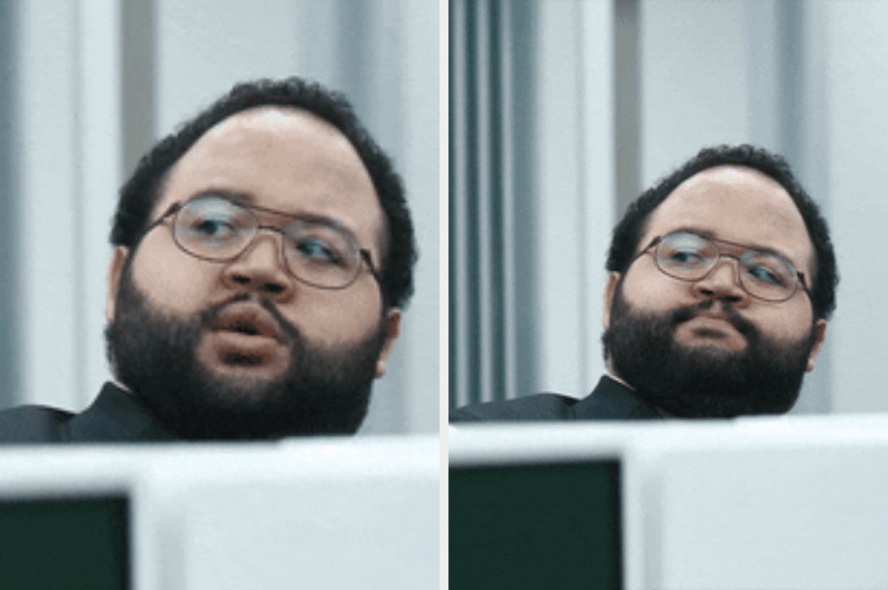 Man in business attire peers over a cubicle in an office setting, expressing curiosity or eavesdropping