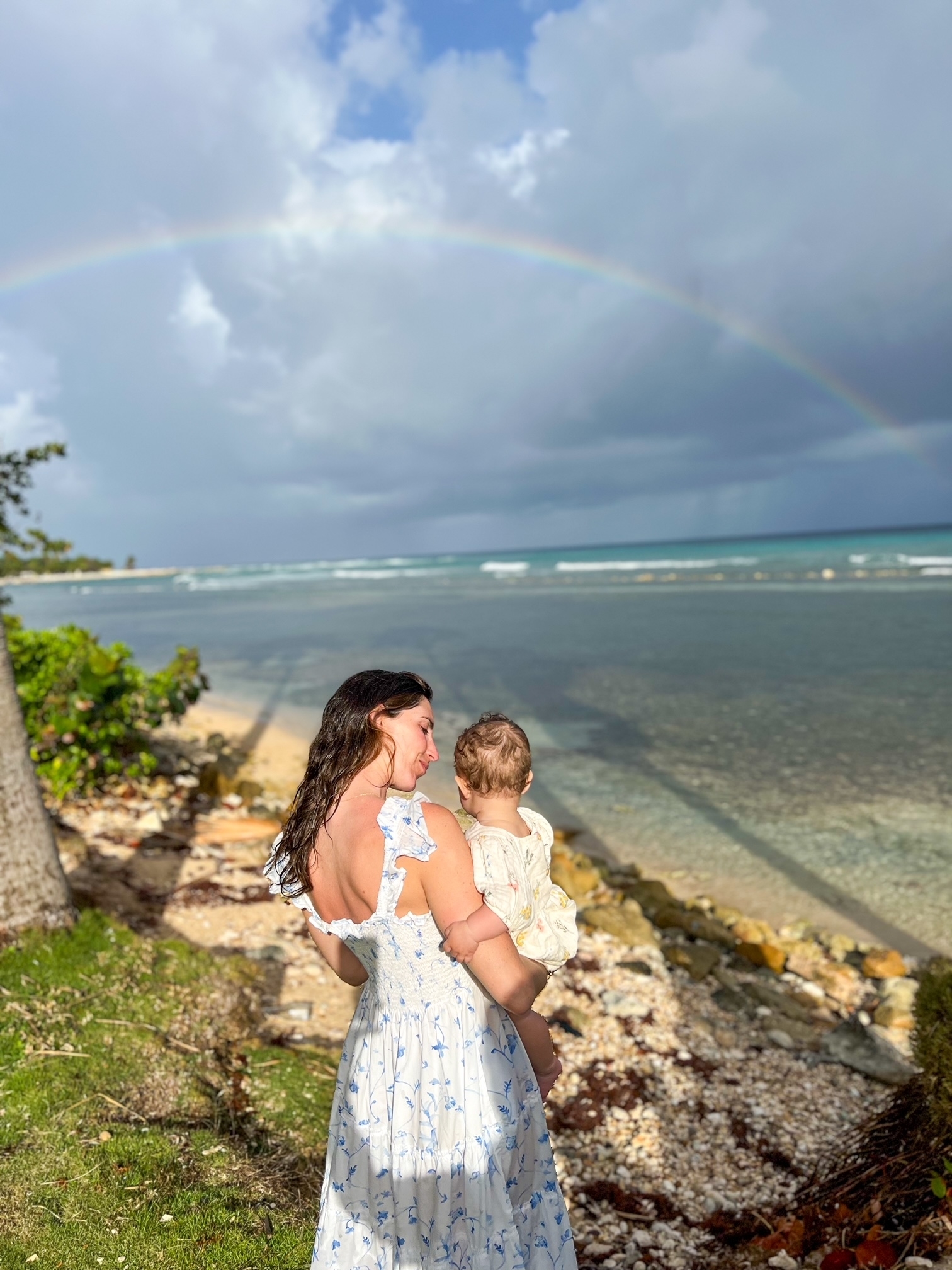 Woman holding a child, both looking at a rainbow over a beach landscape