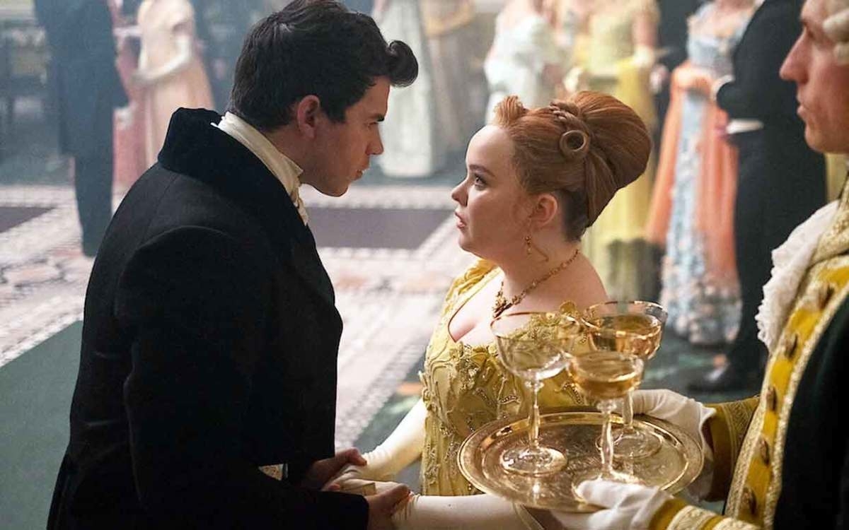 Dos personajes de la serie Bridgerton interactúan, uno sostiene copas, vestidos con trajes de época