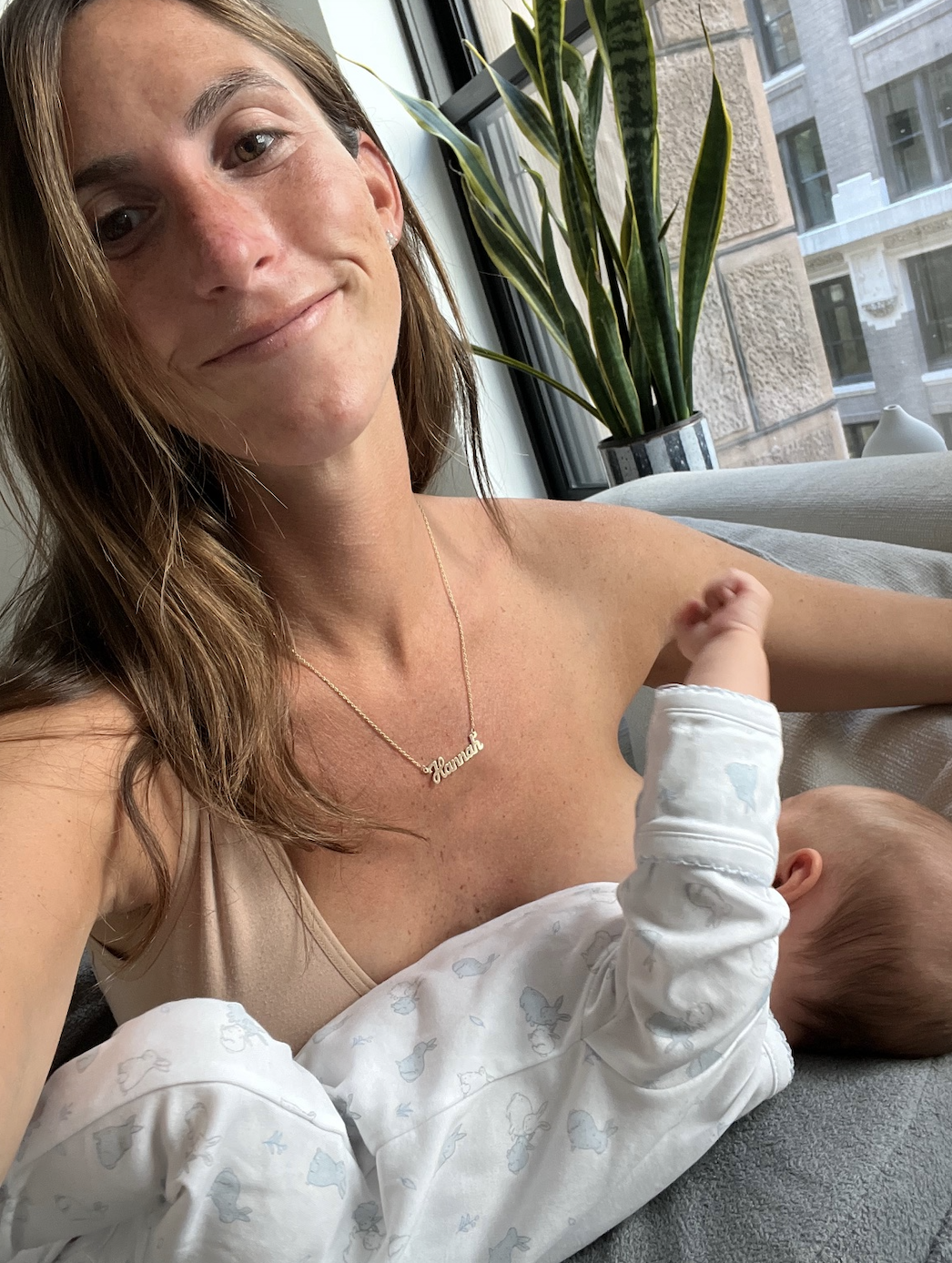 Woman smiling at camera holding a sleeping baby in a onesie, with "Jessica" necklace, indoors by a window
