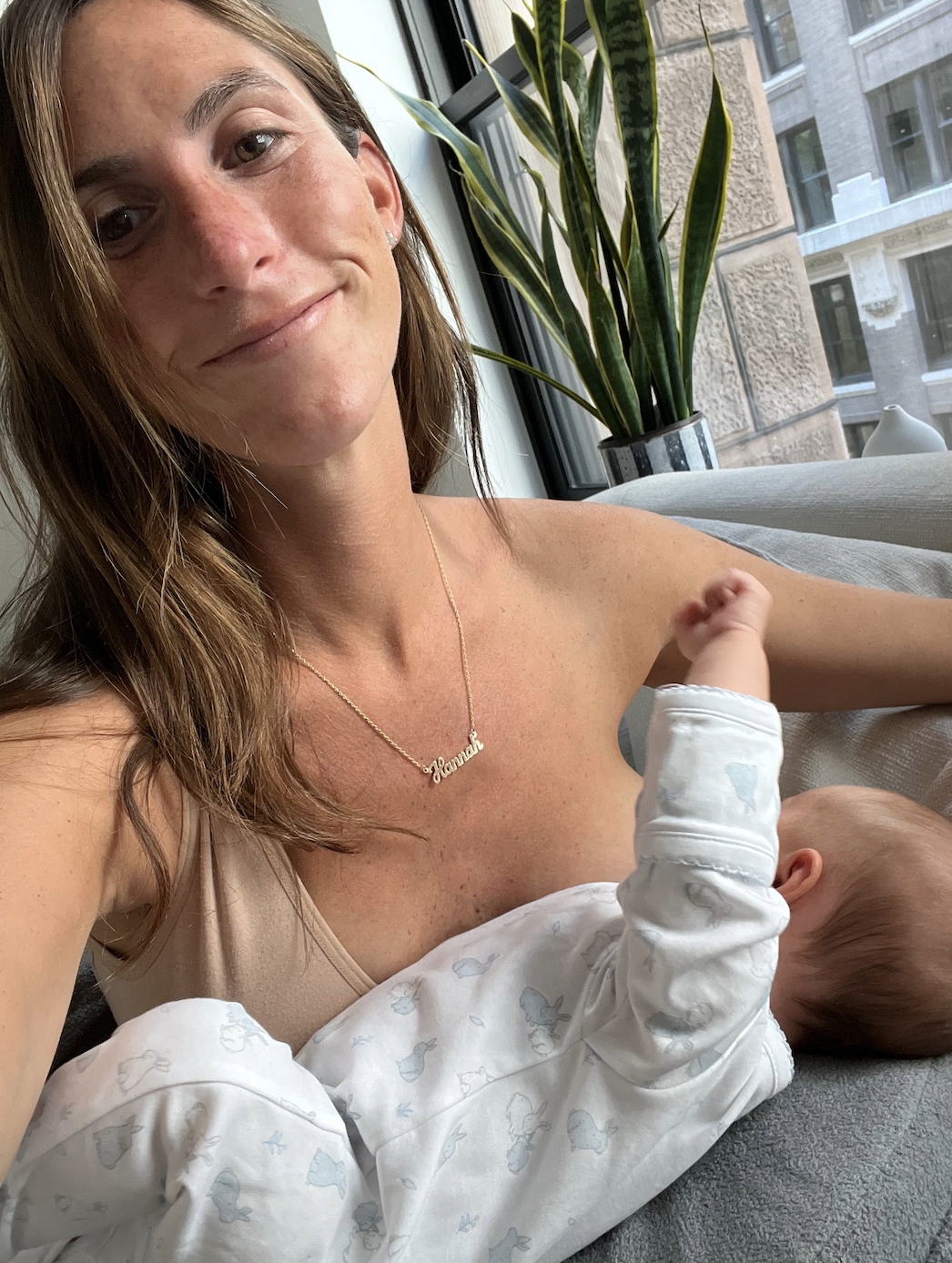 Woman smiling at camera holding a sleeping baby in a onesie, with &quot;Jessica&quot; necklace, indoors by a window