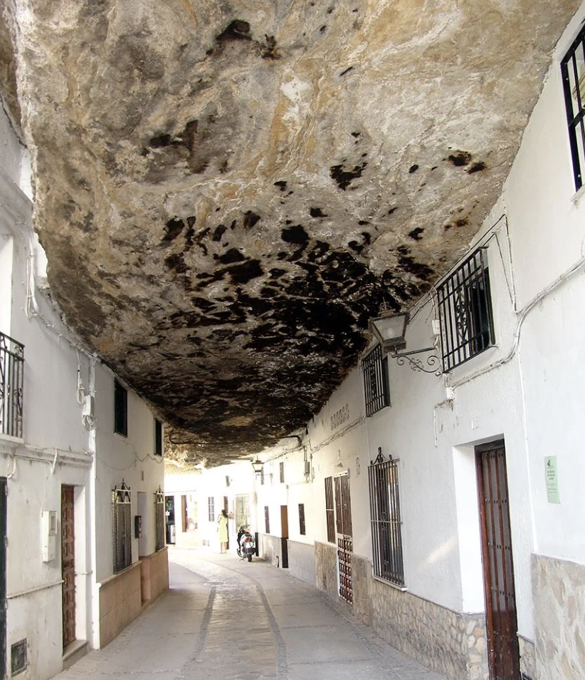 Narrow street with buildings under a large overhanging rock formation
