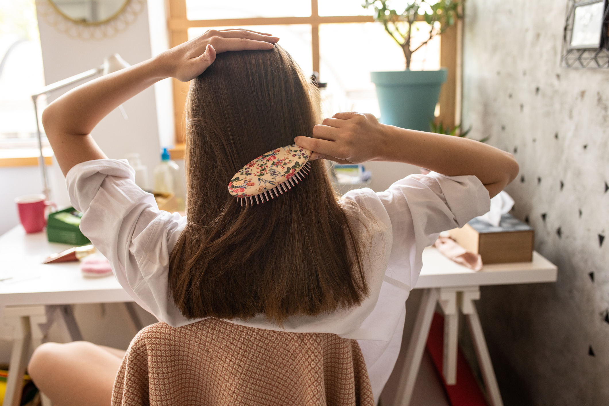 Person combing hair with a brush, seated at a desk, facing away from the camera