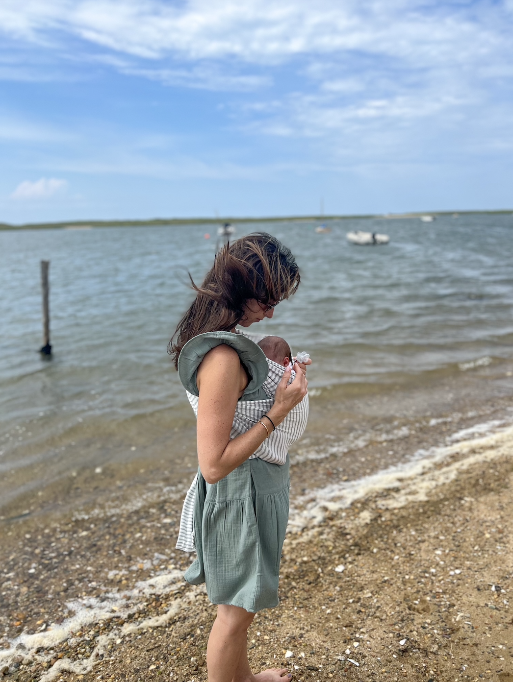 Woman holds baby, both facing the sea. She's in a sleeveless dress; the baby in a onesie. They stand on a sandy beach