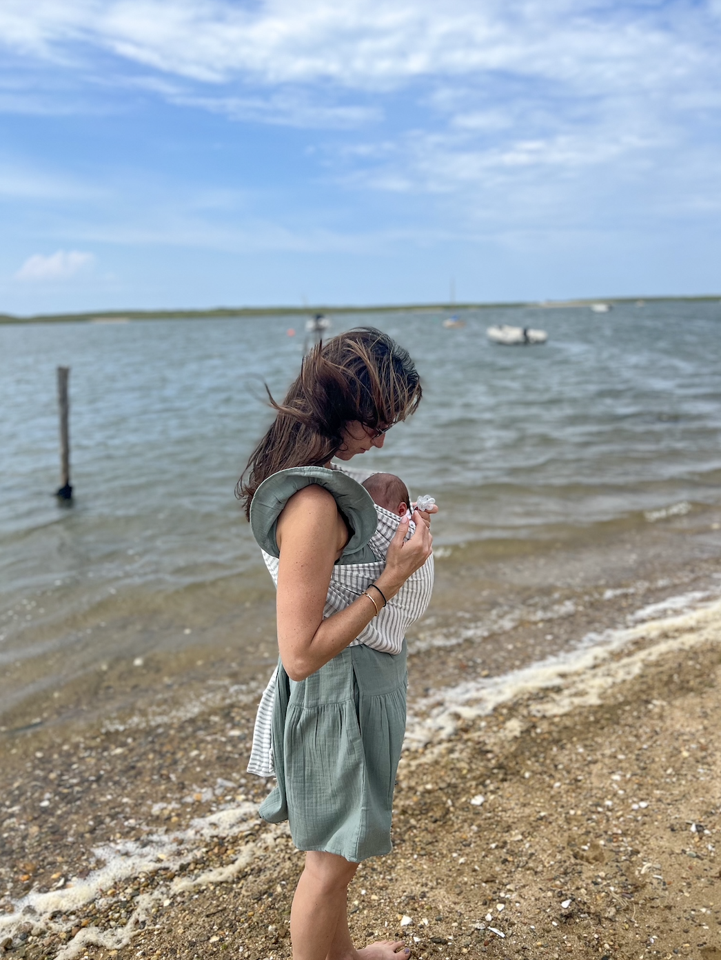 Woman holds baby, both facing the sea. She&#x27;s in a sleeveless dress; the baby in a onesie. They stand on a sandy beach