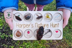 Child holding a wooden 'Harper's Nature Hunt' board with collected natural items in labeled compartments