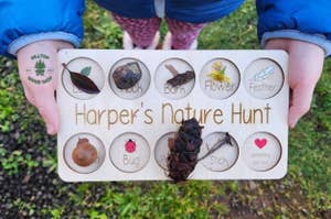 Child holding a wooden 'Harper's Nature Hunt' board with collected natural items in labeled compartments