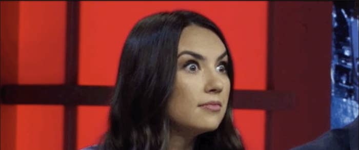 Famke Janssen with a surprised expression on a TV show set with red background