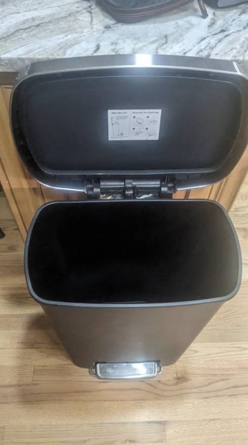 Stainless steel trash can with a foot pedal and lid open, against a wood floor